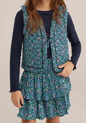 Pige iført en blå langærmet top under en blomstret quiltet vest og matchende lagdelt blomster nederdel med grønne, hvide og blå blomster.