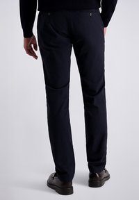 Marineblauwe broek met een gestructureerde stof, voorzien van achterzakken en subtiele stiksels. Gecombineerd met donkere schoenen, gezien vanaf de achterkant.