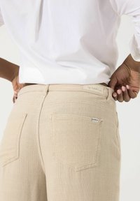 Pantalones texturizados beige con cintura alta, que cuentan con dos bolsillos traseros y una pequeña etiqueta de Garcia en la cinturilla. Combinados con una blusa blanca.