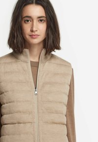 Beige pufferväst med vertikalt quiltat mönster, hel dragkedja fram och hög krage. Bärs över en brun tröja med mjuk textur.
