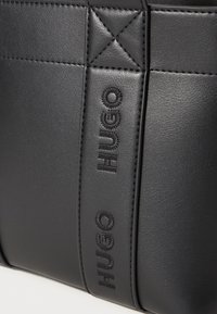 Borsa in finta pelle nera con logo "HUGO" impresso, caratterizzata da una texture trapuntata e dettagli cuciti per un tocco in più.