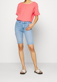 Top in lino rosa chiaro con maniche corte abbinato a pantaloni Bermuda in denim azzurro e sandali rosa chiaro con una fibbia decorativa.