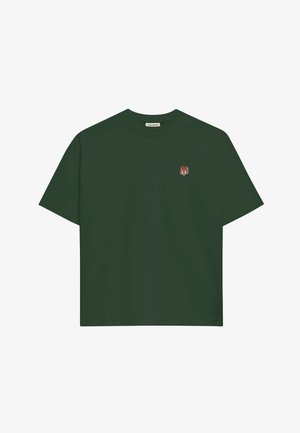 Jonge man draagt een donkergroen T-shirt met een klein oranje logo en een witte en grijze honkbalpet met de tekst "Prohibited Sporting Goods Inc.".