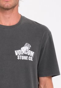 Camiseta de algodón gris con cuello redondo y mangas cortas. En la parte frontal presenta un texto gráfico en blanco y un diseño de logo.