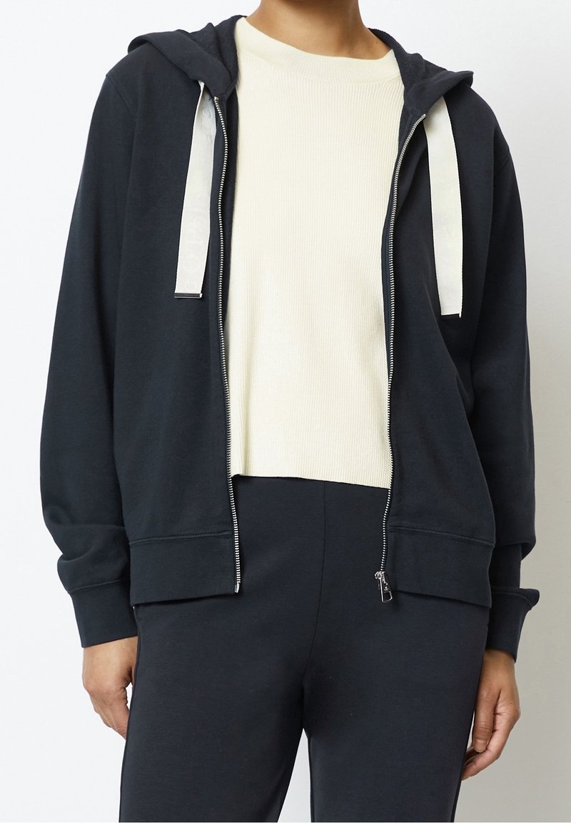 Personne portant un sweat à capuche noir zippé avec des cordons blancs par-dessus un t-shirt blanc côtelé à col rond et un pantalon noir.