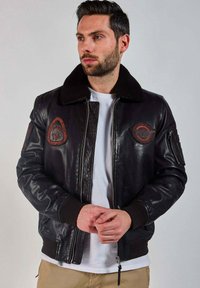 Homme portant une veste en cuir noir avec des patchs, un t-shirt blanc et un pantalon beige, se tenant devant un fond gris uni.