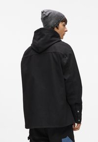 Melnas kapuces jakas, izgatavotas no kanvas auduma, ar pogām uz piedurknēm un brīvu piegriezumu. Valkāta ar pelēku adītu cepuri.