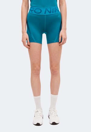 Modèle portant un short de cyclisme Nike bleu sarcelle, des chaussettes blanches et des baskets blanches sur un fond uni.