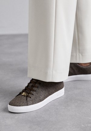 Brown textured low-top sneakers with white soles and gold lace charm, worn with off-white straight pants on gray floor.