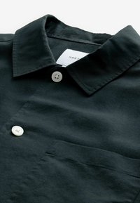 Chemise noire à boutons blancs, col et poche poitrine visible, montrée en gros plan et soigneusement pliée.