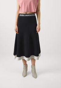 Svart skjørt med ribbet hvit kant og grå elastisk midjebånd med "KARL LAGERFELD" branding; paret med spisse beige ankelstøvler.