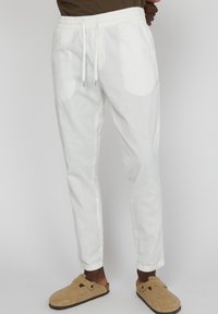 Pantalones de lino blancos con cintura elástica y cordón. Corte ajustado y tobillos con puño. Combinados con zapatos de ante marrón tipo slip-on.