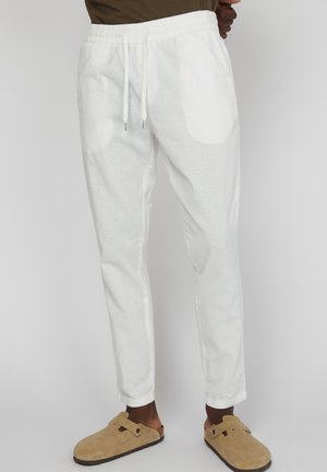 Pantalons en lin blancs avec une taille élastique et un cordon de serrage. Coupe fuselée et poignets. Associés à des chaussures à enfiler en daim marron.