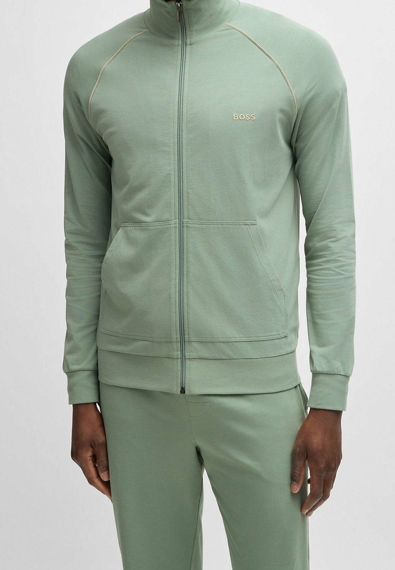 Groene jas met rits en hoge kraag, voorzien van een voorrits, twee zijzakken en lichtgekleurde biezen. Voorzien van een logodetail op de borst.