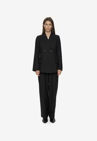Zwarte op maat gemaakte dubbel-gebreide blazer met zijzakken, gecombineerd met losse zwarte broek en puntige zwarte schoenen, op een neutrale achtergrond.