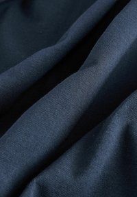 Tummansininen kangas, jossa on sileä, tekstuurinen pinta, ja hienovaraisia ryppyjä ja taitoksia, mikä viittaa pehmeään ja joustavaan materiaaliin.