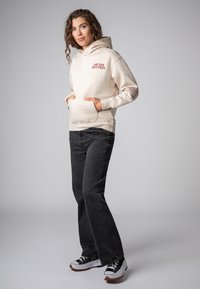 Beige hoodie met de tekst "Love Has Many Faces", voorzak en geribbelde manchetten, gecombineerd met donkergrijze flared jeans en sneakers.