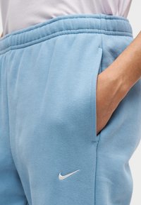 Ръка на човек в джоба на светлосини потни панталони Nike с ластична талия и бяло лого Swoosh на лявото бедро