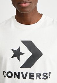 T-shirt bianco in cotone con un grande logo nero che presenta una freccia e una stella, insieme alla parola "CONVERSE" in lettere nere in grassetto.