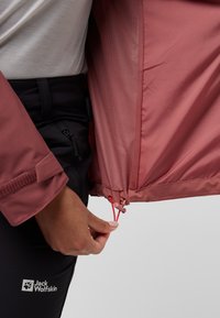 Giacca rosa leggera con un esterno liscio, fodera in rete e orlo regolabile con cordini rossi. Pantaloni neri con logo visibile.
