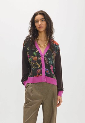 Mujer con un cárdigan negro con patrones florales y de aves coloridos, ribete púrpura y pantalones marrones de talle alto, de pie con las manos en los bolsillos.