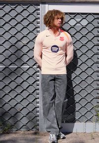 Light pink long-sleeve jersey with a collar, featuring a circular Spotify logo and team crest, paired with faded gray jeans and sneakers.