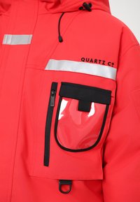 Rote wasserdichte Jacke mit reflektierenden silbernen Streifen, einer schwarzen Reißverschluss-Tasche und einer durchsichtigen Patch-Tasche. Adjustable Kapuze inklusive.