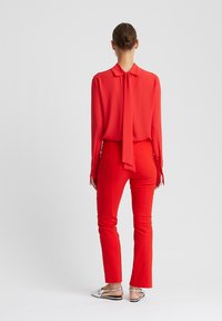 Rode blouse met een strik bij de kraag, gecombineerd met rode nauwsluitende broek. Gladde textuur; model draagt metallic instapschoenen.