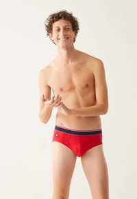 Culotte en coton rouge avec une taille rayée bleu marine et un petit logo sur le côté gauche. Texture lisse et design ajusté.