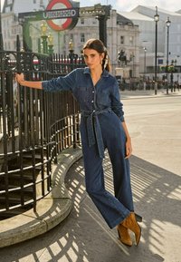 Denim kombinezon z dolgimi rokavi, z gumbi spredaj, dvema žepoma na prsih in tkanim pasom. V kombinaciji s bež visokimi škornji. Urbani ozadje.