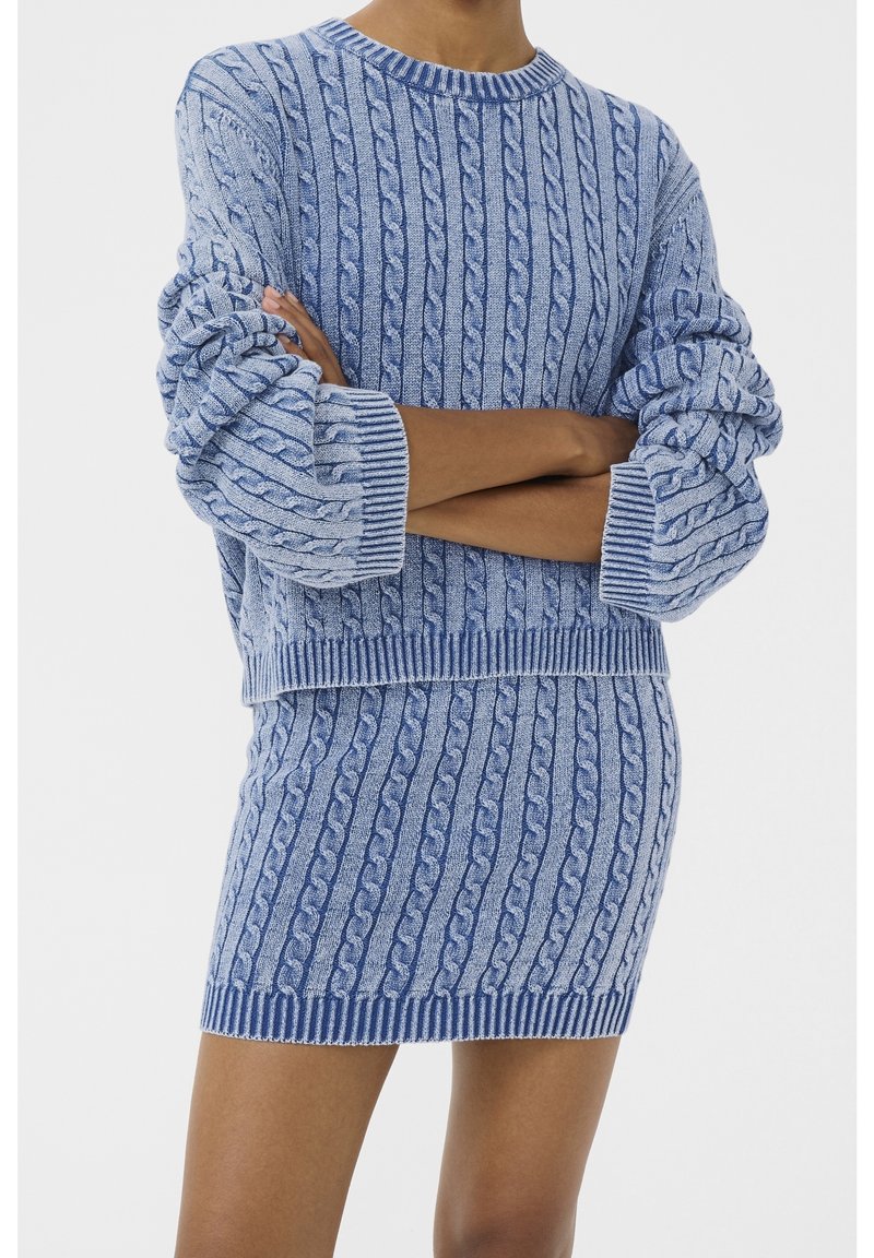 Personne portant un pull tricoté en bleu avec un motif à torsades et une jupe assortie, les bras croisés, montrée des épaules jusqu'à mi-cuisse sur un fond uni.