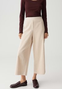 Vrouw in romige, wijde, textuur-pantalons, gecombineerd met een donkerbordeaux lange mouw top en bijpassende bordeaux loafers op een witte achtergrond.