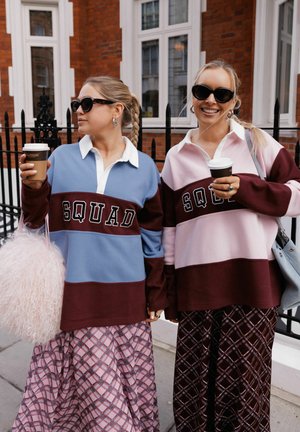 Due donne che indossano felpe color-block con la scritta "SQUAD", gonne a fantasia, occhiali da sole, tengono tazze di caffè e sono in piedi su un marciapiede in città accanto a una recinzione nera.