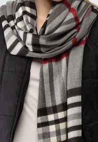 Femme portant une veste noire et une grosse écharpe à carreaux gris, noir, blanc et rouge enroulée autour du cou.