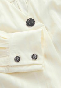 Chemise jaune clair avec des rayures verticales, boutons ronds noirs et détails de manches avec plis et accents de couture.
