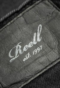 Black leather label with embossed text "Reell est. 1997," set against a textured black fabric background; minimalistic design, no additional patterns.