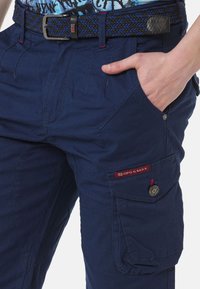 Marine cargobroek van een mix van katoen en polyester. Kenmerkt zich door meerdere zakken, rode accenten, en een gestructureerde zwart-blauwe gevlochten riem.