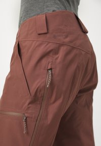 Marron vandringssøm med letvægtsmateriale, der har lynlåslommer og en glat tekstur. Inkluderer et Core-Tex-logo på taljebåndet.
