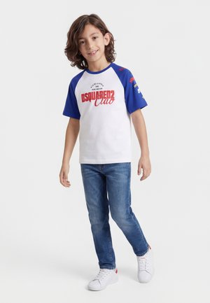 Dreng med skulderlangt hår iført en hvid og blå grafisk t-shirt, blå jeans og hvide sneakers, stående på en ensfarvet baggrund.