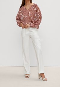 Lichtroze blouse met bloemenpatroon, ruime pasvorm en v-hals. Gepaard met witte, rechte broek en lichtbeige hoge sandalen.
