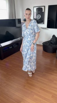 Άτομο με μπλε φόρεμα με λουλούδια και λευκά σανδάλια, που στέκεται σε ξύλινο πάτωμα σε ένα καθιστικό με τηλεόραση, πουφ και κορνιζαρισμένα έργα τέχνης.