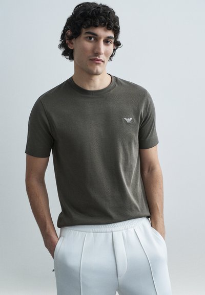 Donkergroene olijfgroene T-shirt met korte mouwen en een ronde hals, met een klein zilveren logo op de linkerborst. Gladde, aansluitende textuur.
