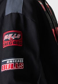 Black jacket featuring embroidered Chicago Bulls logos in red and white. Textured fabric with grey accents on the shoulder. Zipper closure.