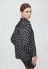 Blusa nera con grandi pois bianchi, maniche lunghe, polsini con bottoni e colletto. Tessuto leggero con una vestibilità rilassata e dettaglio a piega sul retro.