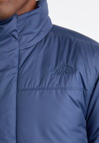 Dark blue insulated jacket with a high collar, featuring a subtle embroidered logo on the chest and a smooth, matte texture.