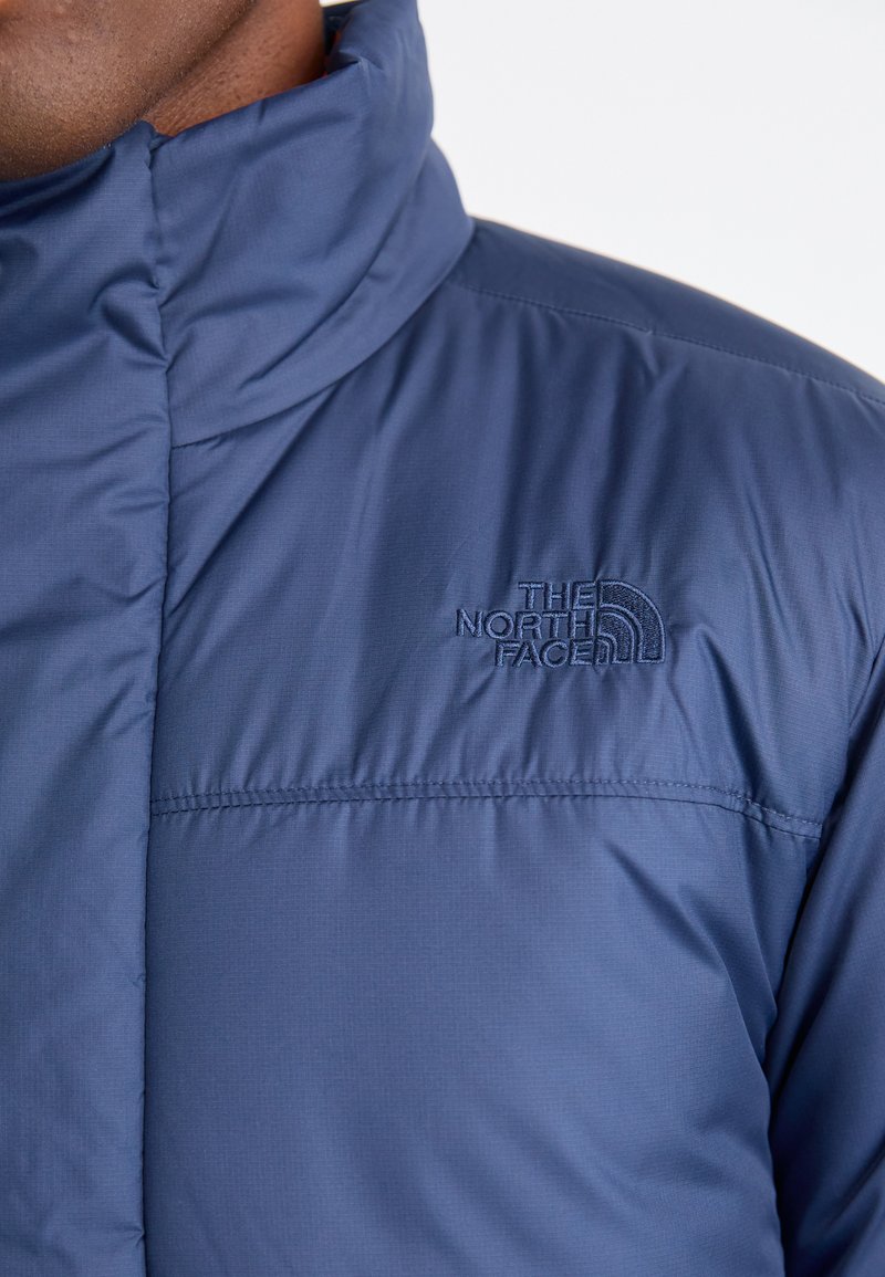 Giacca isolante blu scuro con colletto alto, caratterizzata da un sottile logo ricamato sul petto e una superficie liscia e opaca.