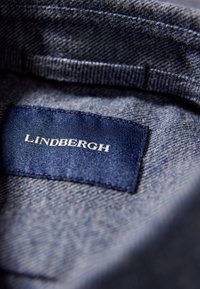 Denim stof met een close-up van een marineblauwe label met witte tekst "Lindbergh." Zichtbare stiksels langs de randen.