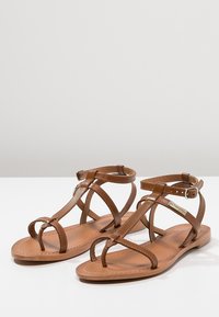 Bruine leren sandalen met meerdere geïnterweven banden en een enkelgesp. Platte zool en minimalistisch ontwerp. Glad oppervlak, geen patronen.