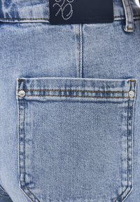 Lichtblauwe denim spijkerbroek achterzak met zichtbare stiksels, twee metalen klinknagels en een zwart leren merkpatch met een ontwerp van strass-steentjes erboven.