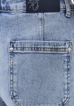 Tasca posteriore di jeans in denim azzurro chiaro con cuciture visibili, due rivetti metallici e una targhetta in pelle nera del marchio con un design di strass sopra.